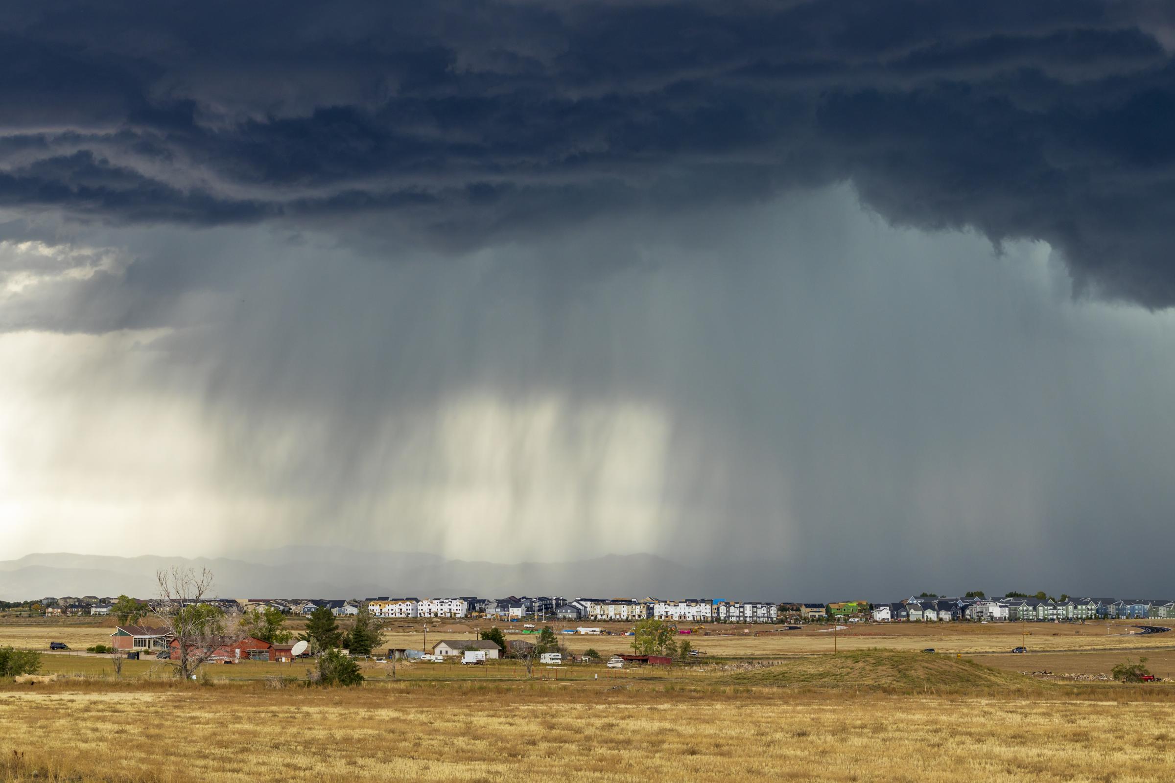Rainfall from dark clouds over a suburban neighborhood, with dry fields in the foreground.