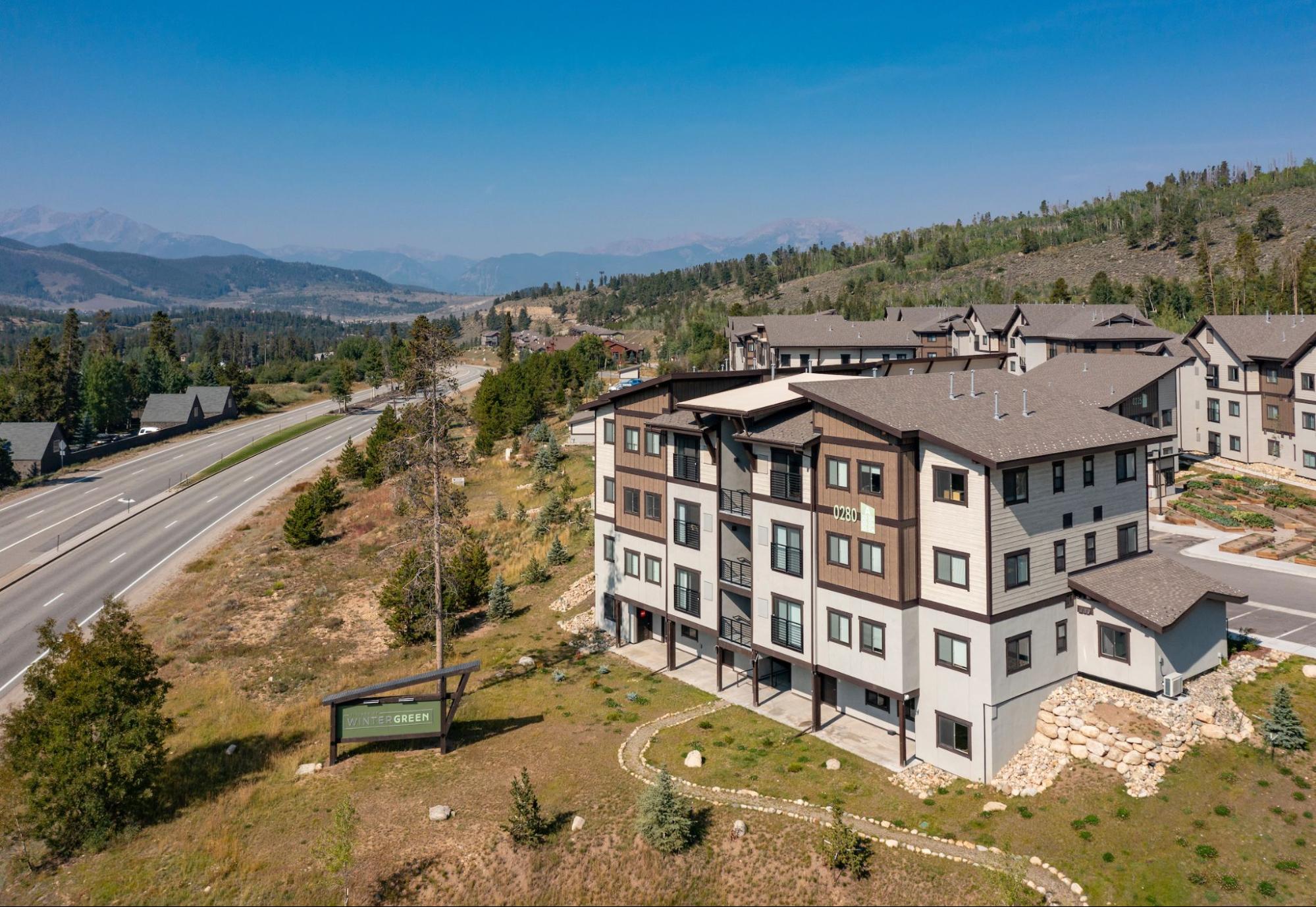 Apartment buildings with wood and stucco siding near a highway, surrounded by trees and mountains in the background. A sign in the foreground reads “Winter Green.”