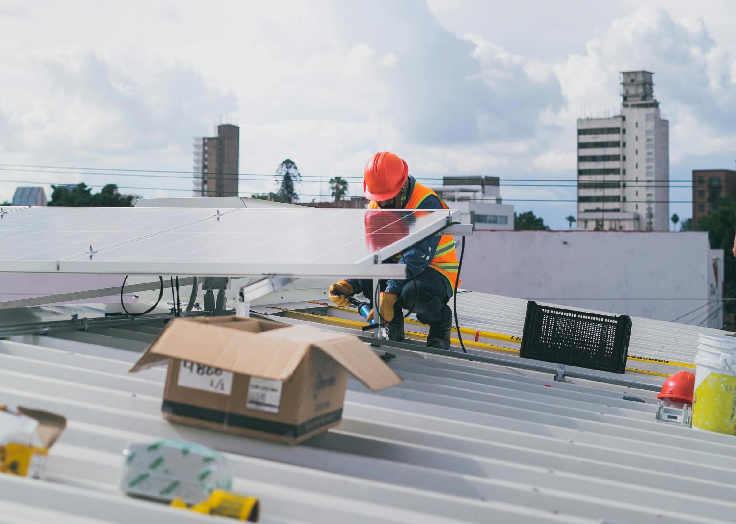Worker in a hard hat and safety vest installing a solar panel on a metal rooftop in an urban area.